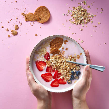 Womans hands holding a bowl of organic yogurt smoothie with strawberries, banana, blueberry, oat flakes and biscuits on pink background. Flat lay.の写真素材