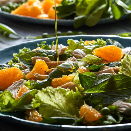A stream of olive oil pours into the prepared salad with natural green vegetables, chicken pieces, orange slices in a blue ceramic plate. Close-up.の写真素材