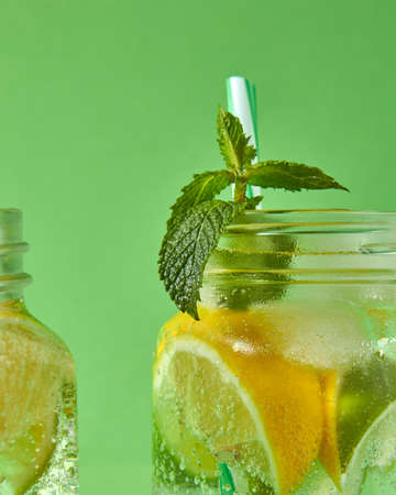 A sprig of green mint in focus in a glass jar with a refreshing natural handmade cocktail. Citrus fruits slices of lemon and lime, ice, water and plastic straw in the glass.の写真素材