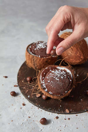 A woman decorates a homemade chocolate ice cream in a coconut with chocolate balls on a board on a...