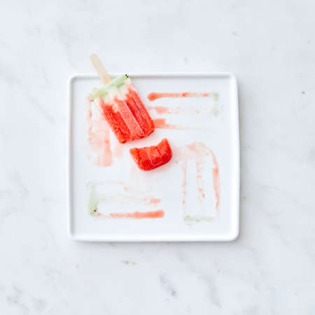 Broken fruit ice cream lolly on a white plate with a pattern from the thawing ice cream on a gray...