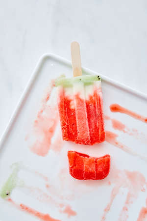 Frozen juice on a stick. Broken ice cream in a plate with splashes of ice cream on a gray marble...