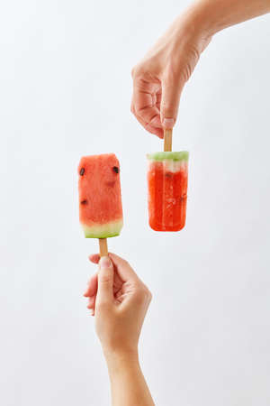 A man's hand holds a slice of watermelon on a stick, in the form of an ice cream, and a red ice...