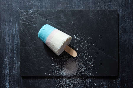 Homemade white blue ice cream lolly decorated with coconut chips on a black slate on a dark concrete...