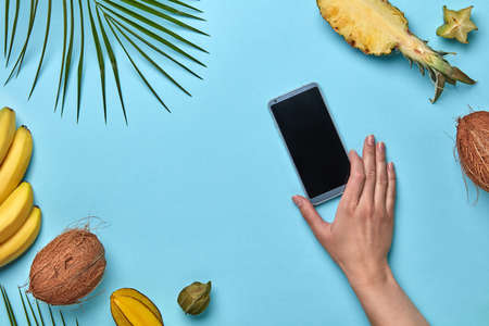 A womans hand holds a mobile phone against a blue background around a tropical fresh fruit with copy space. The concept of buying online. Flat layの写真素材