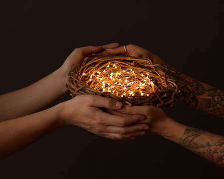 Mens hands and womens hands with a tattoo holding a nest of branches with yellow garlands around a dark background.の写真素材