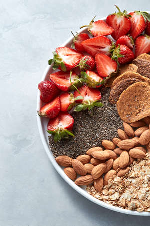 Homemade oat granola with almond nuts, slices of strawberries, seed chia, in a white plate on a gray...