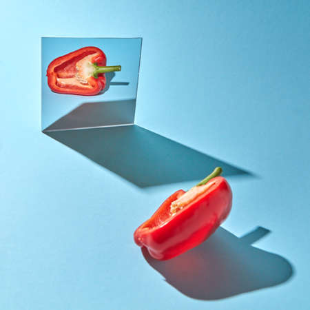 Half a ripe pepper is reflected in a mirror on a blue background with copy space and shadows. Healthy vegetableの写真素材