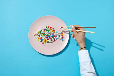 Fish from colorful pills and tablets on a white plate and womans hand with chopsticks on a blue background with copy space. Flat layの写真素材