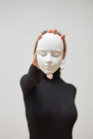 Plaster mask in the hand of young female instead face on a white background, place for text. Concept The masks we wear.の写真素材