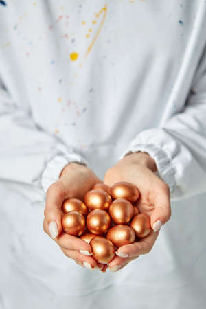 The hands of a young girl hold golden painted eggs on the background of an apron with paint brigs. Wealth conceptの写真素材