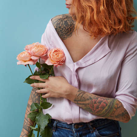 A young woman with a tattoo holds in her hand a bouquet of pink roses around a blue background with copy space. Mothers day conceptの写真素材