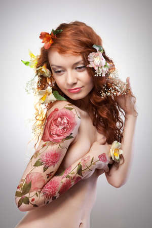 Beautiful red-haired girl with tattoo adorned with flowers on a gray background with copy space. Spring conceptの写真素材