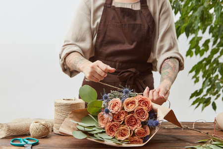 Florist workplace - womans hands are arranging a new bouquet with roses and decorative green leaves on a wooden table, process step by step. Place for text. Mothers Day.の写真素材