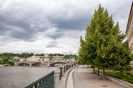 Urban scene with green riverfront along river with view to bridge and historical buildings of Prague...