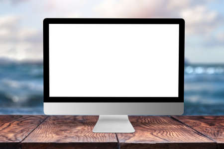 White blank mock up computer display on a wooden table against blurred seascape natural background with bokeh, copy space. Working remotely concept.の写真素材