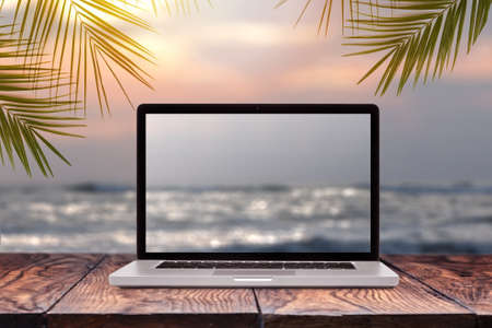 Notebook monitor with blurred seascape sunrise picture on a wooden table against the same background with green palm leaves frame, copy space. Working at sea, outside office concept.の写真素材