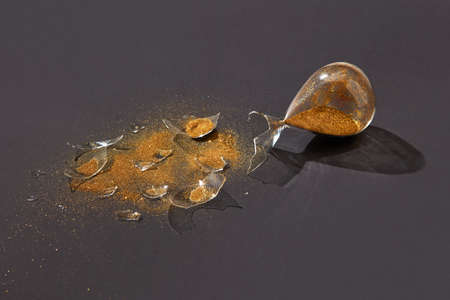 Old-fashioned aged crashed sandglass with glass shatters and golden sand on a black background with hard shadows, copy space.の写真素材
