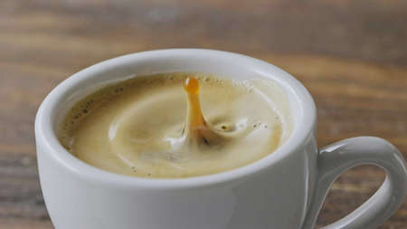 Close-up coffee splashes fall into a cup of espresso with froth on a brown wooden table. Slow motion, Full HD video, 240fps, 1080p.の写真素材