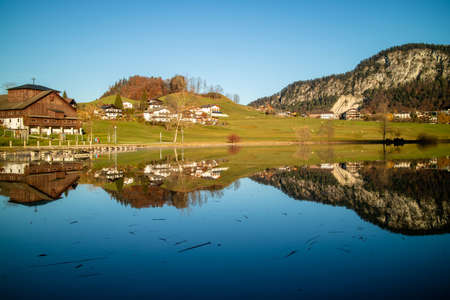 Beautiful countryside landscape with traditional houses, natural hills and lake with reflection of it in blue clean water on a background of clear sky.の写真素材