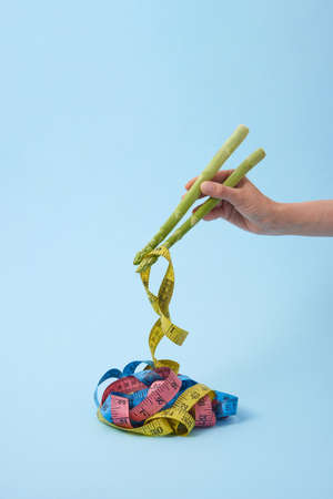 Woman's hand with asparagus sticks is taking measure tapes as a colorful noodle against pastel blue background, copy space. Colored measuring tapes as Japanese or Chinese food.の写真素材