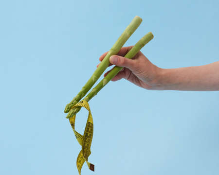 Female's hand with asparagus sticks is taking measure tape as a noodle against pastel blue background, copy space. Yellow measuring tape as Japanese or Chinese food.の写真素材