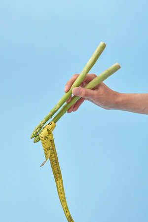 Asparagus spears in a woman's hand holding yellow measuring tape as a noodle against pastel blue background, copy space. Yellow measuring tape as Japanese or Chinese food.の写真素材