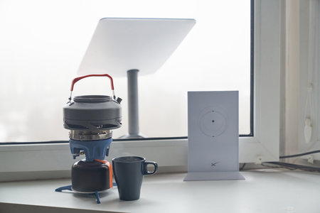 KYIV, UKRAINE - JANUARY 24, 2023: Close-up of Starlink antenna and wi-fi router on windowsill with portable burner stove with kettle and cup of tea beside.のeditorial素材