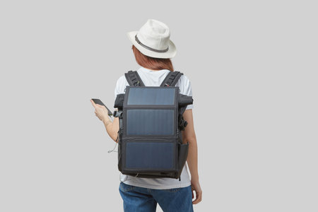 Girl carrying backpack with flexible solar panel attached to smartphone in her hands, isolated on light grayの写真素材