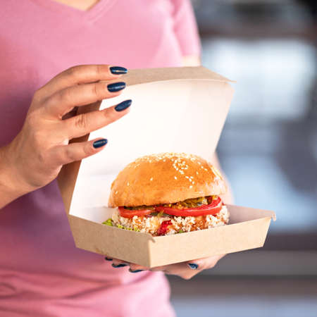 Food delivery concept. Closeup of Female hands holding opened paper box or lunchbox with Fresh burger. Nutritious Fast food snack or lunch. Square format for posting on social media.の写真素材