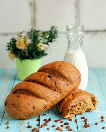 Loaf of wheat bread with raisins and milk bottle on kitchen table. Light breakfast and flower pot on blurred background. Morning food. Rustic still life concept. Vertical format.の写真素材
