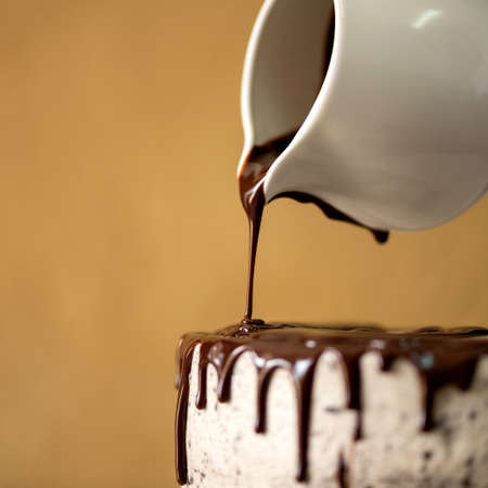 Birthday cake cooking. Hot melted chocolate is poured from jug on top of white cake, Close up. Mustard color background. Sweets for holiday event. Blank for confectionery advertising banner.の写真素材