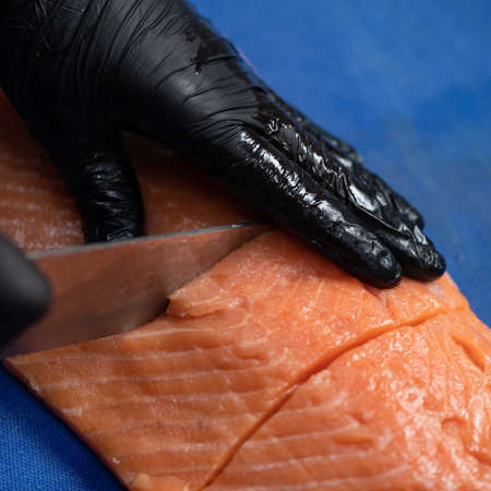 Chef prepares raw red fish. Hands in black glove cutting pieces of fresh salmon fillet with big...
