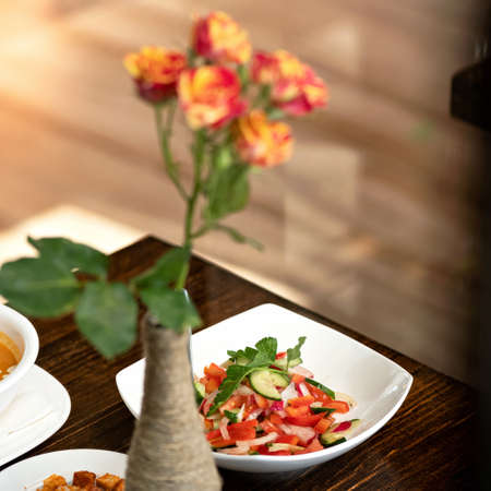 Colorful salad from fresh vegetables and greens on table. Holiday event concept. Vase with rose flower on blurred foreground, soft focus.の写真素材