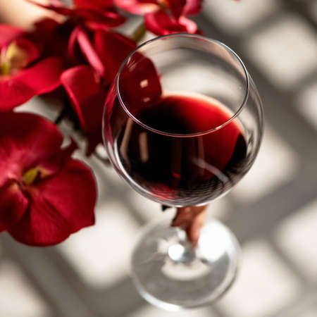 Glass of red wine and red orchid flowers on wooden table with sun glare. Alcoholic drink. Close up shot, soft focus.の写真素材