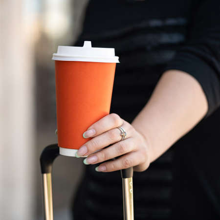 Blank of Disposable Paper Coffee cup in female hand. Woman with suitcase holding Paper cup of coffee. Close up shot. Mockup or template cardboard container for drinks.の写真素材