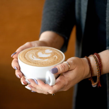 Cup of creamy cappuccino coffee in female hands. Woman holds portion of hot aromatic drink with froth. Square format, soft focus. Close up shot. Copy space.の写真素材