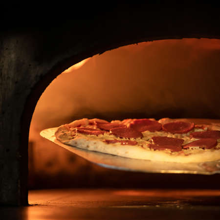 Hot pizza on wood freshly baked. Ready to eat pepperoni pizza with salami against blurred background of furnace with burning fire. Authentic Italian Restaurant or pizzeria. Soft Focus.の写真素材