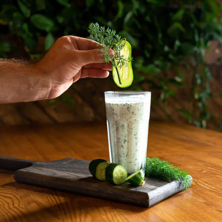 Chef creates refreshing vitamin smoothie drink with mixed greens and vegetables. Male hand adds sprig of dill and slice of cucumber in glass with dairy cocktail. Healthy power of nature. Soft focus.の写真素材