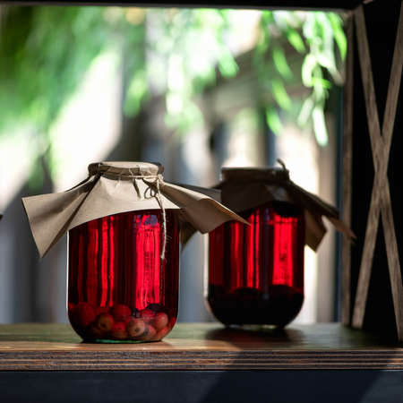 Red berry compote in glass jars. Two cans of preserved or canned cherry drink on wooden table in...