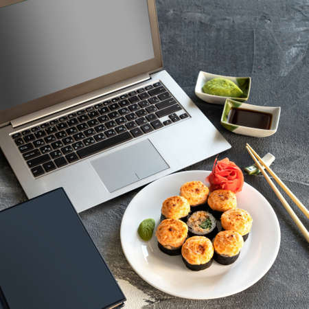 Japanese rolls and business, sushi break. Laptop computer with blank screen, diary, plate Hot baked Sushi roll, sauces and chopsticks on workplace. Healthy Oriental food. Top Side view. Soft focus.の写真素材