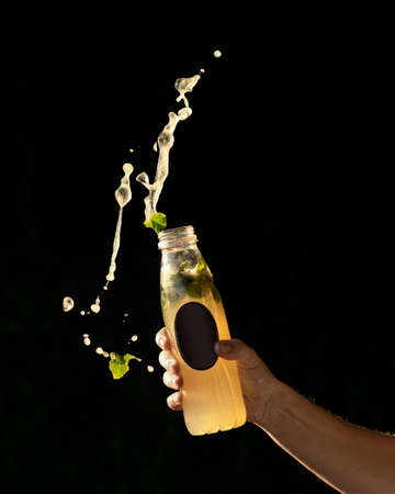 Splashing of bottle drink. Orange refreshing beverage with mint splash from transparent bottle with blank label. Male hand holds plastic container on black background. Template for branding.の写真素材