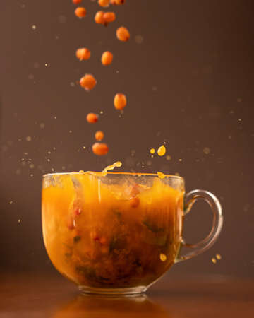 Sea buckthorn tea. Berries falling into glass cup with Orange herbal drink. Close up shot in motion...