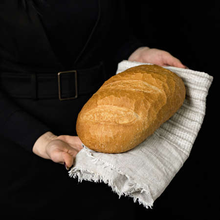 Golden wheat bread on linen cloth in female hands. Woman holding fresh whole loaf on black...