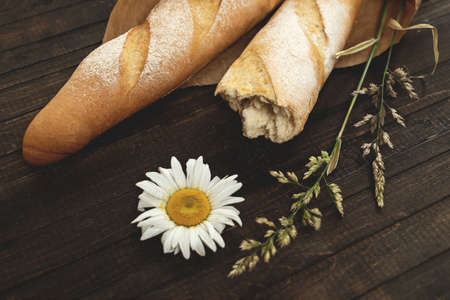 Golden wheat baguettes. Two craft loaves of crispy bread on brown wooden table with wildflowers on it. Top view. Horizontal format.の写真素材