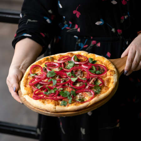 Woman holds baked homemade pizza. Female hands holding wooden cutting board with sliced pizza with red onion, meat, parsley and melted cheese. Close-up shot. Soft focus. Copy space.の写真素材