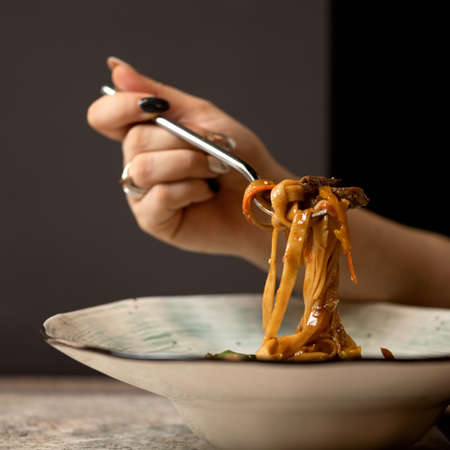 Woman eating hot pasta with fork. Italian food, noodles or spaghetti with tomato sauce on blured background. Food plate and female hand. Side view. Close-up shot. Soft focus.の写真素材
