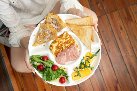Plate with breakfast in female hands on wooden background. Toasts, egg omelette, crepes, cheese and ham slices, arugula, lettuce and cherry tomatoes on serving platter. Top view. Horizontal format.の写真素材