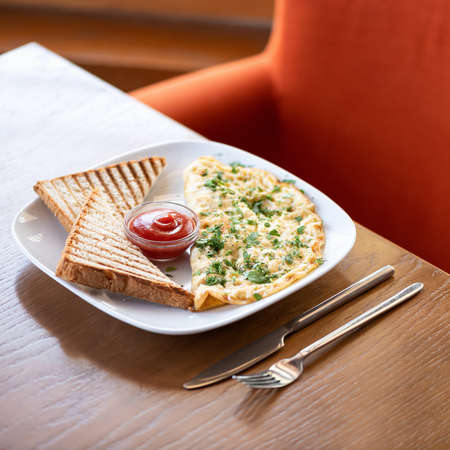 Eggs scrambled omelet with herbs toasts and tomato ketchup. Plate with food and cutlery on wooden restaurant table. Red armchair on blurred background. Soft focus. Copy space.の写真素材