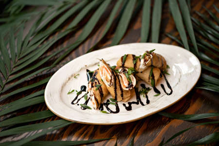 Platter of thin pancakes with red fish filling and soy sauce topping. Festive Snack on table decorated with palm leaves. Horizontal format. Soft focus.の写真素材